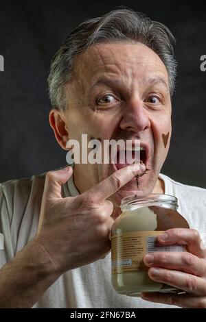 A middle-aged man greedily eats some chocolate cream with his finger straight from the glass jar making a very funny expression Stock Photo