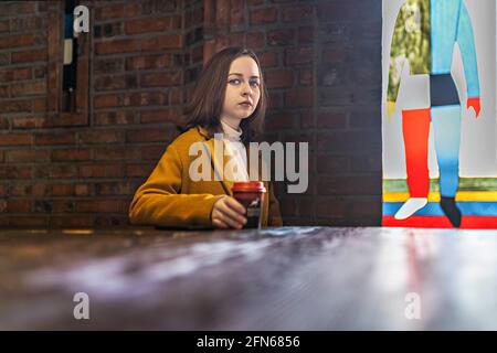 Young girl in a cafe, with a cup of coffee waiting for a meeting.In the background a brick wall and a stained glass colored window. Loft. Stock Photo