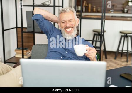 Senior cheerful man enjoys morning coffee sitting at the workplace, mature businessman using laptop indoor, grey haired male holds cup and looks at the computer monitor, rests on the chair Stock Photo