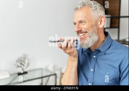 Senior cheerful man sending voice message, using smartphone app for recording and recognizing voice commands, mature grey haired businessman holds phone and talks in the mic, making audio notes Stock Photo