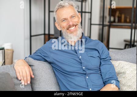 Charismatic grey haired man in smart casual wear sits on the couch and looks at the camera. Portrait of handsome mature guy with wide toothy smile. Health and wellbeing concept Stock Photo