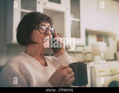 Senior woman holding cup of coffee and talking on mobile phone in kitchen at home Stock Photo