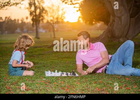 Son laying on grass and playing chess with father. Little boy play chess with parent. Cognitive development. Intellectual kids game. Smart children. Stock Photo