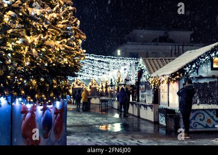 Festive Christmas and New Year's fair square in the city at night, falling snow, New Year tree and houses with garlands. Stock Photo