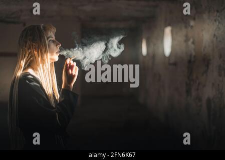 Blond young woman smoking a cigarette in ruined room with light shinning from window Stock Photo