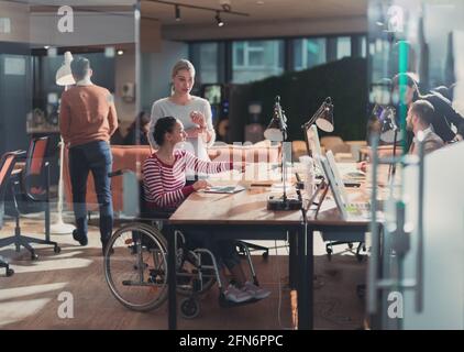 Disabled businesswoman in a wheelchair in modern coworking office space. Colleagues in background. Disability and handicap concept. Selective focus Stock Photo