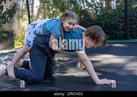 Lynwood, Washington, USA.   Four yeard old girl trying to climb on her teenage brother's back on the trampoline.  (MR) Stock Photo