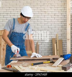 Asian carpenter with a hard hat and dustproof glasses use a sander machine to smooth plywood surface by abrasion with sandpaper. Morning work atmosphe Stock Photo
