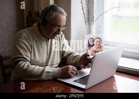 Smiling senior man wear earphones wave to camera having video call on laptop, happy elderly male in headphones sit on couch at home talk using modern Stock Photo