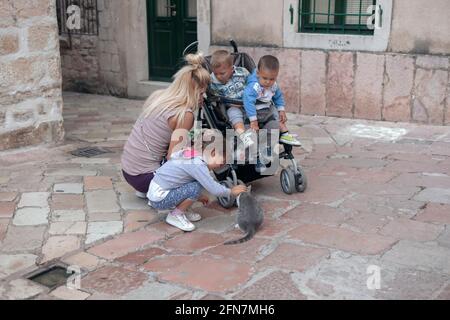 Montenegro, Sep 22, 2019: Mother and baby brothers watching little girl playing with pussycat on the street of Kotor Old Town Stock Photo