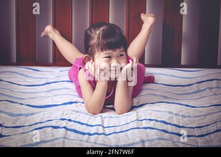 Adorable little asian girl in a purple dress smiling. Cute child  lying barefoot on bed in bedroom. Vintage picture style. Stock Photo