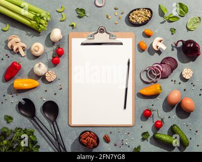 Food ingredients, salad serving utensils and clipboard with white paper sheet. Various of vegetarian cooking ingredients on gray background. Diet menu Stock Photo