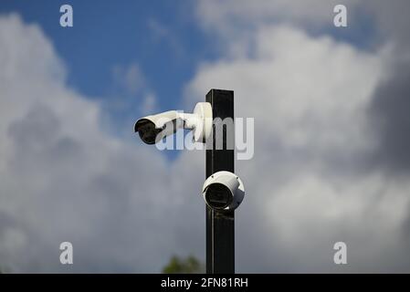 Two white security cameras mounted on a black pole, covered in cobwebs, with clouds and blue sky in the background Stock Photo