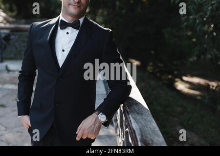 A man in a classic black suit with a bow tie and a watch on his arm leans on a handrail against a background of trees. Place for your text Stock Photo