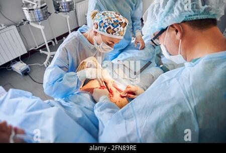 Medical workers in surgical suits performing plastical surgical operation. Plastic surgeon and assistant wearing medical face masks and sterile gloves while removing excess fat from patient abdomen Stock Photo