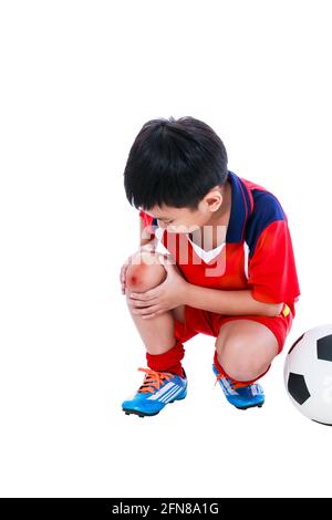 Sports injure. Youth asian soccer player with football in red uniform injured at knee. Isolated on white background. Studio shot. Full body. Stock Photo