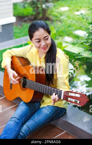 Pretty musician smiling and having fun enjoy hobby concept. Asian woman playing classic guitar at backyard, outdoor with bright sunlight. Positive hum Stock Photo