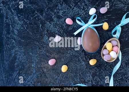 chocolate egg with blue bunny ear bows and multicolored chocolate eggs on a black textured background. top view Stock Photo