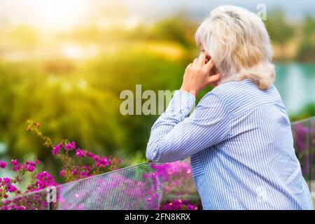 Woman chatting on her mobile phone on an elevated patio outdoors overlooking greenery and the river below in the warm glow of the rising sun Stock Photo