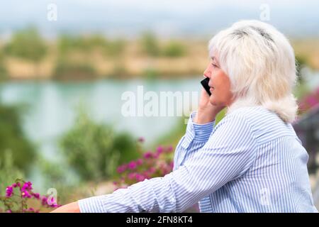 Woman standing on an outdoor balcony chatting on her mobile phone with a quiet smile as she overlooks a lake below in close up with copyspace Stock Photo