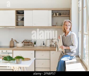 Thoughtful beautiful mature woman looking in window and thinking or dreaming about something, cooking food on kitchen spending free time in morning at home Stock Photo