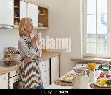 Starting new day. Side view of beautiful thoughtful senior woman drinking coffee or tea and thinking about something, elderly lady in apron enjoying morning while standing in modern kitchen at home Stock Photo