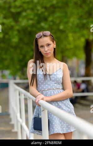 A portrait of a pretty young woman, taken in the summer in a city, leaning against a railing on a nearby river. Stock Photo