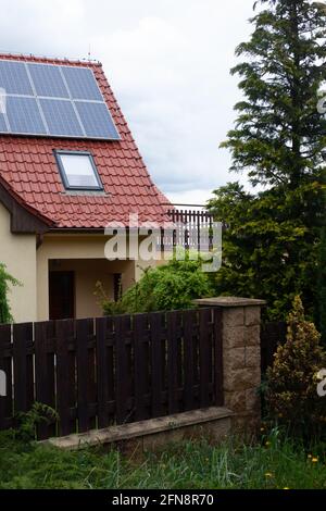 The gable of a house, the roof is covered with solar panels, there is a velux window on the roof. In the foreground is  palisade and a vegetable hedge Stock Photo