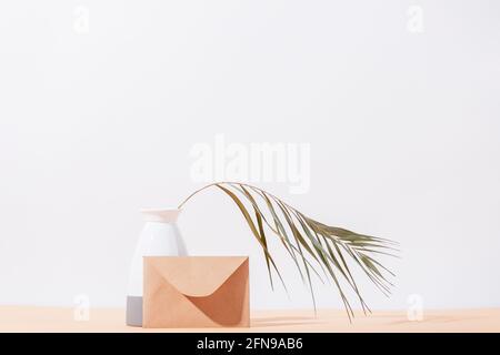 Vase with dry palm leaf and  brown eco paper envelope on beige table against white wall background with empty space. Stock Photo