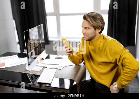 Handsome Caucasian businessman sitting at desk, wear yellow shirt and holding yellow smartphone, Distant worker holding online meeting, using PC Stock Photo