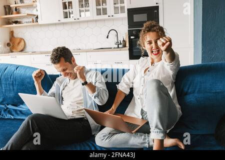 Energetic couple in casual wear sitting on the coach using laptops for online work together. Smiling young freelancers have success in new project Stock Photo
