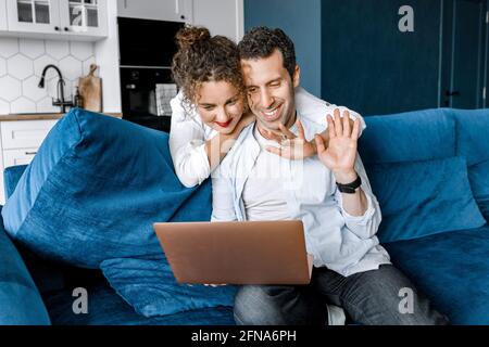 Beautiful young couple cuddle, sit on blue sofa, make video call to friends using laptop webcam. Loving young spouses look at computer screen waving Stock Photo