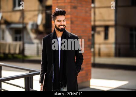 Young indian man wearing demi-season clothes in the street. Young bearded  guy with modern hairstyle with coat, blue jeans and t-shirt against urban  bl Stock Photo - Alamy