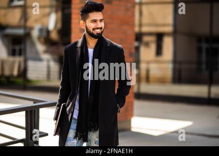 Young indian man wearing demi-season clothes in the street. Young bearded guy with modern hairstyle with coat, blue jeans and t-shirt against urban bl Stock Photo