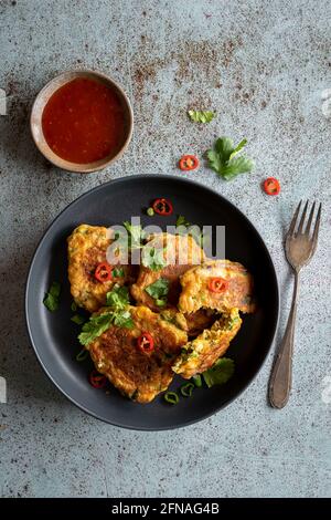 Home made Indonesian corn fritters (Perkedel Jagung or Bakwan Jagung) served with sweet chilli sauce on black plate amd textured blue background, copy