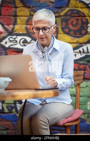 An older woman working on a laptop in a pleasant atmosphere in a bar. Leisure, bar, outdoor Stock Photo