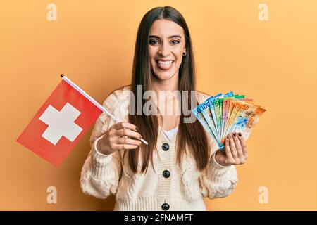 Beautiful brunette young woman holding switzerland flag and franc banknotes sticking tongue out happy with funny expression. Stock Photo