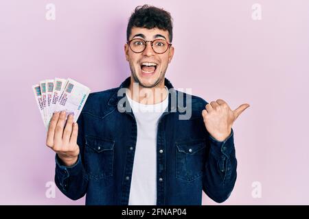 Young hispanic man holding egyptian pounds banknotes pointing thumb up to the side smiling happy with open mouth Stock Photo