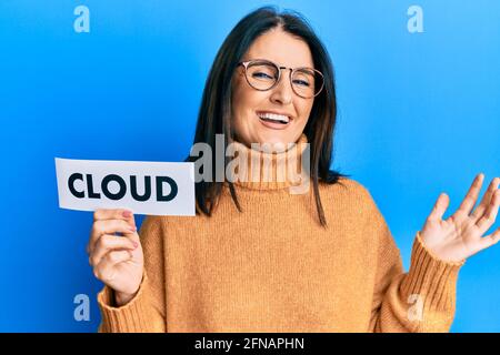 Middle age brunette woman holding paper with cloud word celebrating achievement with happy smile and winner expression with raised hand Stock Photo