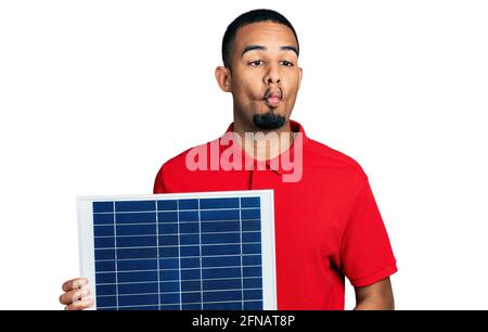 Young african american man holding photovoltaic solar panel making fish face with mouth and squinting eyes, crazy and comical. Stock Photo