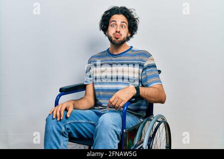 Handsome hispanic man sitting on wheelchair puffing cheeks with funny face. mouth inflated with air, crazy expression. Stock Photo