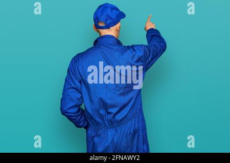 Bald man with beard wearing builder jumpsuit uniform posing backwards pointing ahead with finger hand Stock Photo