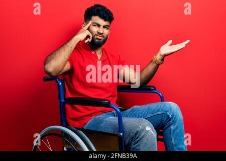 Arab man with beard sitting on wheelchair confused and annoyed with open palm showing copy space and pointing finger to forehead. think about it. Stock Photo