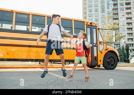 Hard of hearing brothers students with hearing aids in ears jumping near yellow school bus. Inclusion of people with disabilities. Smiling kids going Stock Photo