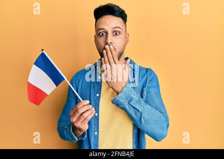 Young arab man holding france flag covering mouth with hand, shocked and afraid for mistake. surprised expression Stock Photo