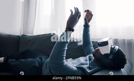 African-American man wearing virtual reality glasses and headset lying on the sofa. Man working using the VR glasses touching something. Focused. High quality photo Stock Photo