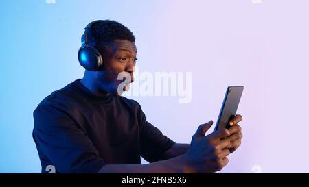 Guy wearing headphones and holding a tablet watching some video. Intense expressions on his face. Blue background. Closeup of a guy using a tablet watching media. . High quality photo Stock Photo