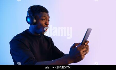 Guy wearing headphones and holding a tablet watching some video. Intense expressions on his face. Blue background. Closeup of a guy using a tablet watching media. . High quality photo Stock Photo