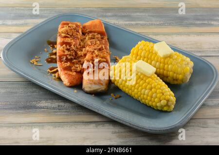 Baked salmon fish fillets with BBQ sauce and crispy onions as topping and corn on the cob with butter on serving plate Stock Photo