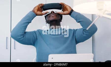 Black man wearing a virtual reality glasses sitting at the table in front of his laptop. Man watching something in the 360 degree virtual environment using the VR glasses. Relaxing. High quality photo Stock Photo